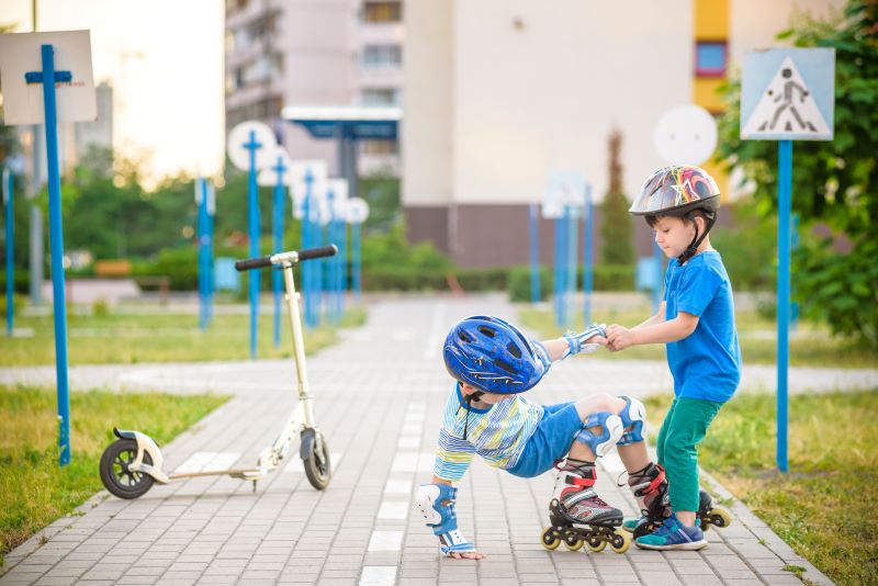 Two children (little boys) in the park, one on the roller skates who has fallen and the other one grabbing his hand trying to lift him.